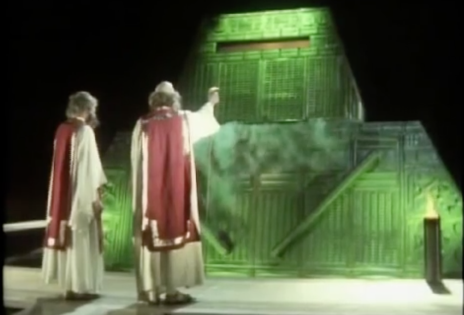 Deep Thought, a giant green supercomputer pouring with steam or smoke from dry ice, towers above 2 philosophers who are asking it a complicated question.